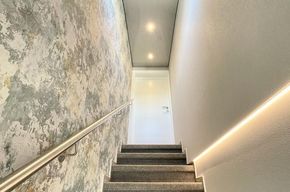 Treppenanlage mit Velourteppichboden und Teppichfußleiste, Vinyltapeten, Spanndecke mit Beleuchtung. Sowie LED-Element an Wand rechts.